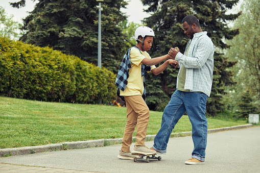 African dad helping his son to stand on skateboard and teaching him to ride outdoors in park