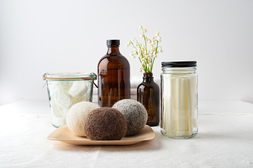 Eco-friendly laundry products: dishwasher tablets in dissolvable wrappers stored in mason jar, liquid laundry soap in refillable bottle, laundry soap sheets in re-useable jar, wool dryer balls on bamboo plate, flower arrangement in re-useable bottle.