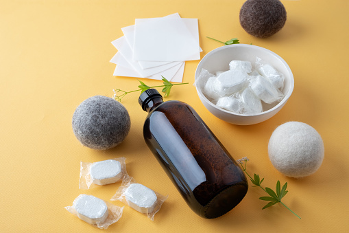 Eco-friendly laundry products: dishwasher tablets in dissolvable wrappers stored in handmade ceramic dish, liquid laundry soap in refillable bottle, laundry soap sheets, wool dryer balls