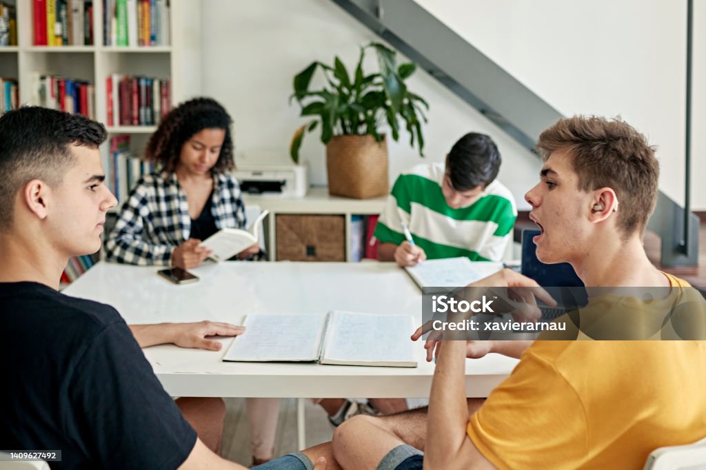 Deaf friends doing homework together Focus on foreground teenage boys in casual clothing sitting at dining table and communicating about their assignment. Part of lifestyle series depicting deaf family and friends. Deafness Stock Photo