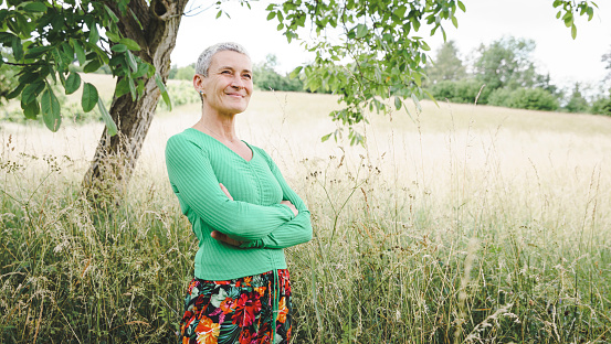 pretty, strong, powerful middle aged woman with green shirt, colorful flower pants, short grey hair posing in green meadow in front of walnut tree