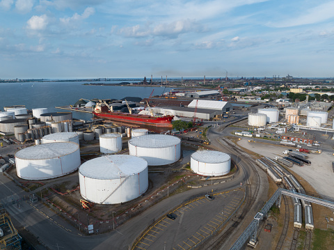 A high-aerial view looking at an oil storage tank field with white petroleum tanks located next to a port. Pictured during the day while a cargo vessel is stationary in the harbor.