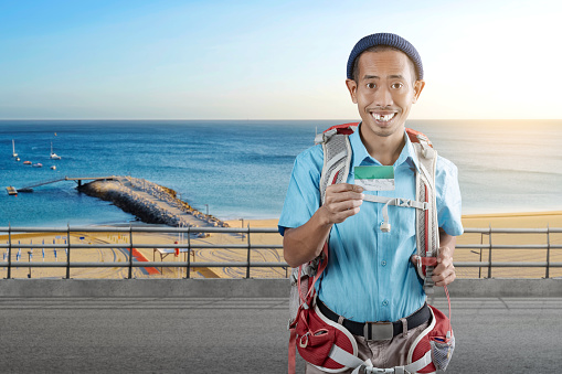 Asian man with a beanie hat and a backpack holding card standing with a beach view and blue sky background