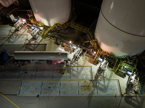 A high-angle aerial view above a large industrial gas facility as a semi-truck with transport trailer is parked, being filled late at night, under bright lights.