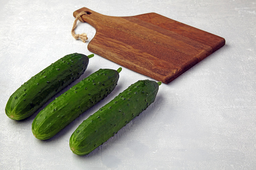 Fresh organic cucumbers and a brown wooden cutting board on a light gray concrete table. Preparing to cook at home