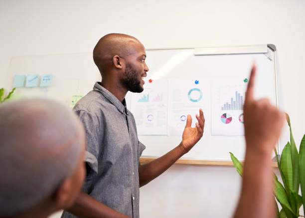 A Black man gives a presentation in the office while a colleague asks a question