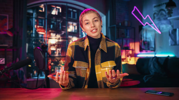 Confident Young Female Talking Into Microphone While Recording Radio Show from Her Loft Apartment. Happy Woman with Short Hair Talking on Camera and Recording Podcast Live on Social Media.