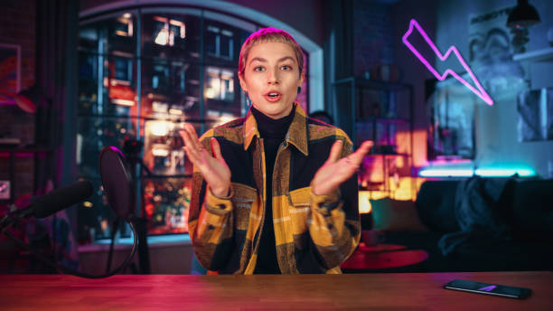 istock Ecstatic Young Female with Short Hair Talking Into Microphone While Recording Podcast from Loft Apartment. Happy Surprised Woman Talking on Camera and Recording Radio Show Live on Social Media. 1409652494