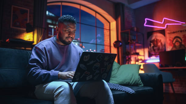 istock Young Stylish Black Man Working from Home on Laptop Computer with Stickers in Cozy Stylish Loft Apartment. Creative Male Smiling, Checking Social Media, Browsing Internet. Flat with Big Windows. 1409652671