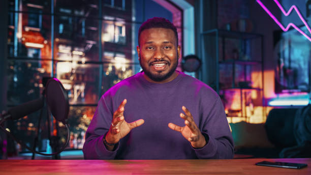 istock Handsome Young African American Man Talking Into Microphone While Recording Radio Show from His Loft Apartment. Happy Black Male Talking on Camera and Recording Podcast Live on Social Media. 1409652795