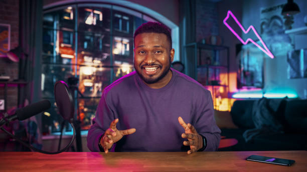 istock Handsome Young African American Man Talking Into Microphone While Recording Radio Show from His Loft Apartment. Happy Black Male Talking on Camera and Recording Podcast Live on Social Media. 1409652806