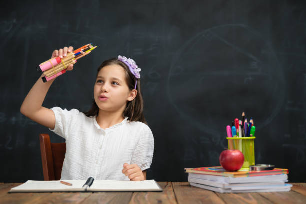 A sweet little elementary school girl sits at a desk working on a writing assignment.  She is dressed casually in a sweater and has a pencil in hand as she looks down to focus on her work.  Her peers can be seen working around her at their own desks as well.