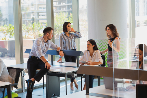 Group of businesspeople talking and working at their desk behind of plexiglass panels between cubicles screen partition and with practicing social distancing for protection against coughs and spitting by separating office tables in a creative luxury office during New normal Covid-19 viruses symptom outbreak at reopen entrance after coronavirus quarantine and lockdown.