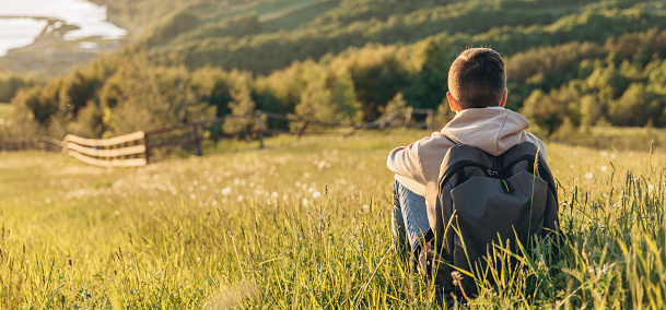Tourist with backpack sitting on top of hill in grass field and enjoying beautiful landscape view. Rear view of teenage boy hiker resting in nature. Active lifestyle. Concept of local travel. Banner