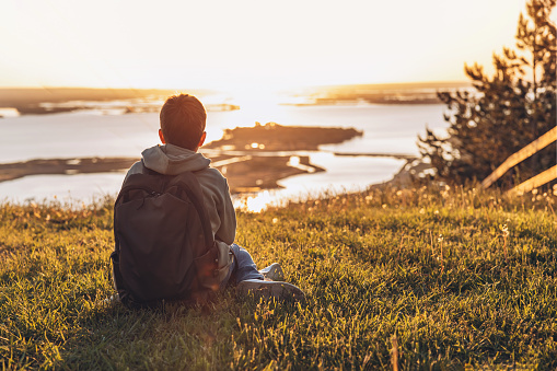 Tourist with backpack sitting on top of hill in grass field and enjoying beautiful landscape view. Rear view of teenage boy hiker resting in nature. Active lifestyle. Concept of local travel