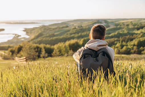 Tourist with backpack sitting on top of hill in grass field and enjoying beautiful landscape view. Rear view of teenage boy hiker resting in nature. Active lifestyle. Concept of local travel