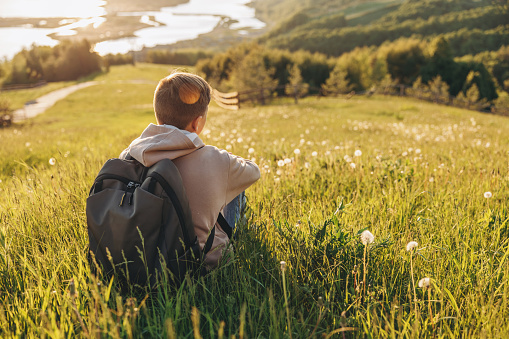 Tourist with backpack sitting on top of hill in grass field and enjoying beautiful landscape view. Rear view of teenage boy hiker resting in nature. Active lifestyle. Concept of local travel