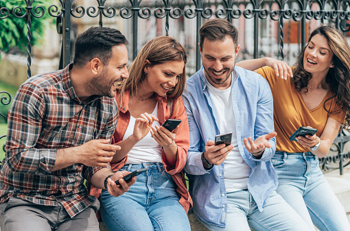 Group of friends with smart phones outdoor. Young beautiful people using smartphones leaning on the fence. Texting in sunny day. Friends surfing the net together.