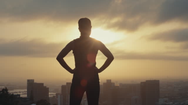 Silhouette of athlete standing in powerful, confident and assertive stance while admiring cityscape at sunset after downtown training workout. Fit, toned and sporty woman feeling ready and successful