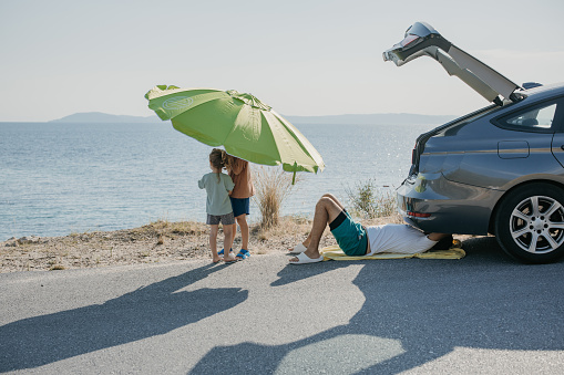 Father lying under a car and trying to fix them on the street, while his son and daughter are looking at the sea.