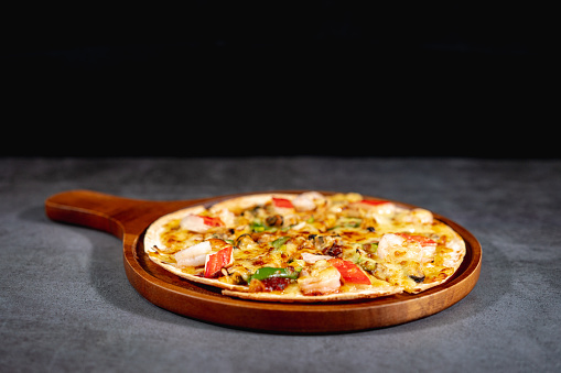 Tasty hot baked seafood crispy pizza - mussels, shrimps and kani with chili pepper and melted mozzarella cheese on round pizza dough, on a wooden pizza pan over the concrete table top.