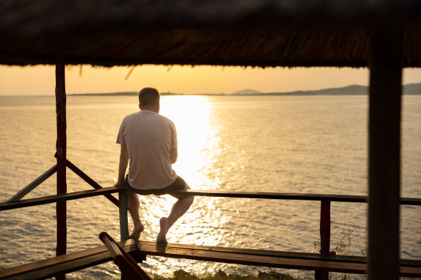 Rear-view of unrecognizable young man, sitting on the fence, while looking at the sunset above the sky