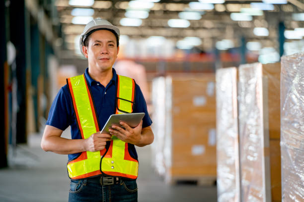 Portrait of Asian warehouse worker or manager hold tablet and look at camer also stand in front of stack of product carton. Concept of professional factory worker support for delivery transport. retrato del trabajador o gerente de almacén asiático sostener la tableta y mirar la cámara también pararse frente a la pila de cartón del producto. concepto de apoyo profesional a los trabajadores de fábrica para el transporte de entrega.
