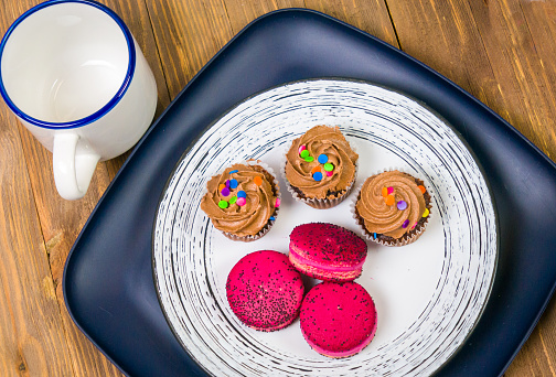 A still life of three chocolate cupcakes and three macarons on a plate with a  cup on a wooden table.