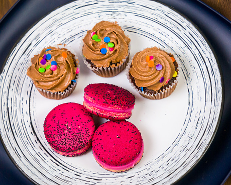 A still life of three chocolate cupcakes and three macarons on a plate  on a wooden table.
