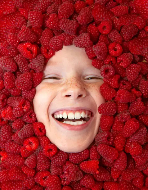Photo of Portrait of a cute laughing child in a frame of ripe juicy raspberries, close-up. The concept of summer, harvest, summer food, life on the farm, childhood, holidays.