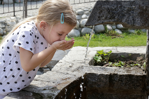 Little girl aged 9 drinking water from a public drinking fountain. Sunny summer day. The girl is wearing blue sun dress.