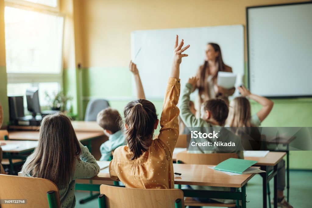 Students raising hands while teacher asking them questions in classroom Education Stock Photo