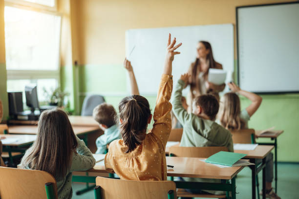 Students raising hands while teacher asking them questions in classroom stock photo