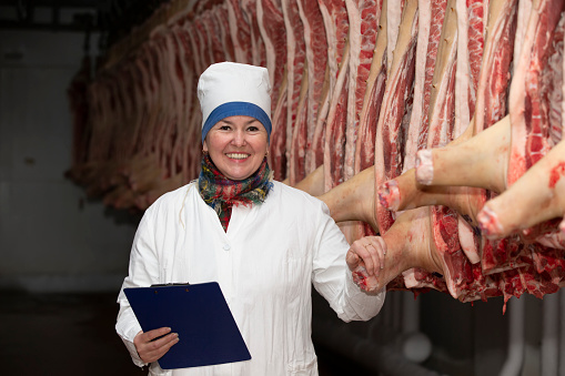 Meat-packing plant worker in front of butchered carcasses. Meat production. A professional butcher between rows of pork carcasses, looking at the camera, posing. A woman works at a meat processing plant.