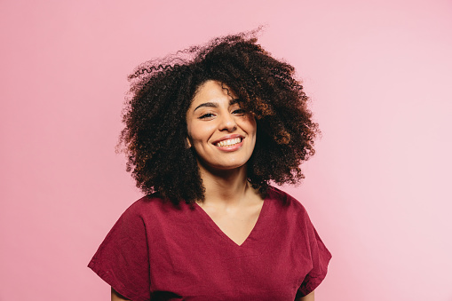 Portrait of a young adult woman with afro hair against a pink background. She's smiling, looking at camera.