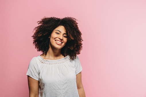 Portrait of a young adult woman with a white shirt against a pink background. Afro haistyle.