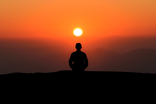 Silhouette of young asian man practices yoga and meditates alone on top of the mountain with beautiful view orange sky and sunrise at morning.
