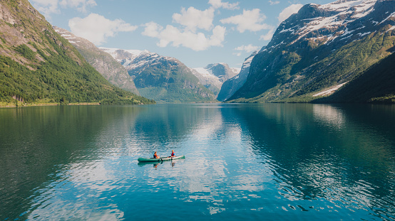 Drone photo of heterosexual couple with their dog sailing in a canoe in the crystal blue glacier lake with view of the snowcapped mountains and the waterfall during sunny day in Vestland County, Western Norway