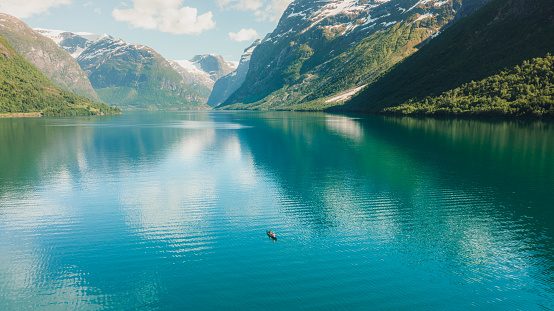 Drone photo of heterosexual couple with their dog sailing in a canoe in the crystal blue glacier lake with view of the snowcapped mountains and the waterfall during sunny day in Vestland County, Western Norway