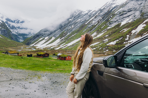 Young happy female with long hair staying by a car looking at the beautiful snowcapped mountains and the old viking village in Norway
