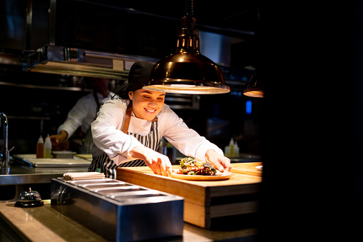 Happy Latin American female chef placing a plate under a hot lamp in a commercial kitchen