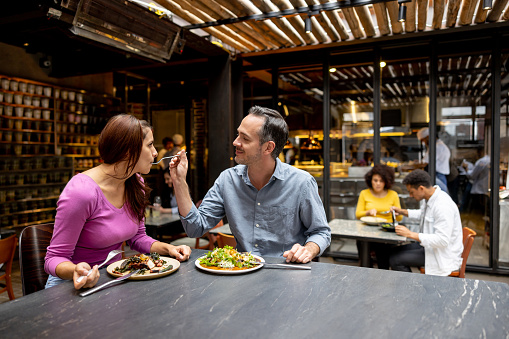 Happy Latin American couple eating together at a restaurant and man giving a taste of his plate to his girlfriend - food service concepts
