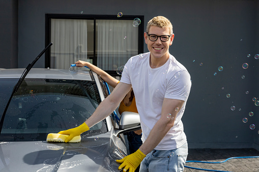 Young Caucasian man is smiling while using sponge and soap water to wash his car in the garage at his home