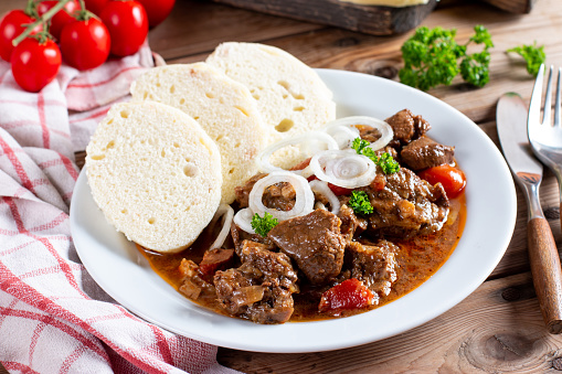 Pork goulash meat with dumplings on white plate, cutlery, garlic, onion, pepper, tablecloth in the background - typical Czech food