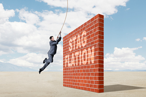 A businessman swings from a rope as he realizes a brick wall of stagflation is dead ahead.