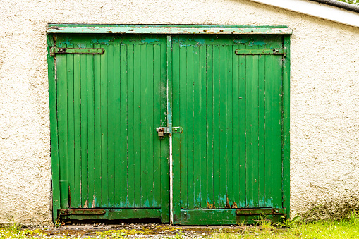 Straight on view of a pair of padlocked green garage doors, with strap hinges, peeling paint and rotten wood