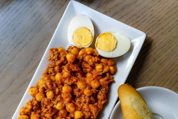 Stock photo showing elevated view of a plate containing a bowl full of a Mediterranean healthy breakfast of shakshouka (shakshuka / chakchouka) served with flatbreads. The recipe consists of chickpeas, egg poached, peppers, onion, olive oil and garlic in savoury tomato sauce spiced with cumin and paprika cayenne pepper.