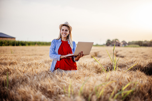 New generation of young woman with organic wheat business, blonde girl on a  field wearing casual clothing and hat, exploring the integrity of organic wheat using laptop