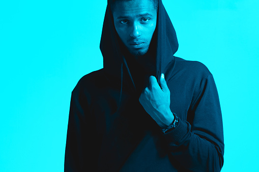 Male portrait of black African-American man at the studio in neon lighting. Color background, copy space. Serious handsome person wear hoodie posing for camera. Photo with smart watch, gadget on hand
