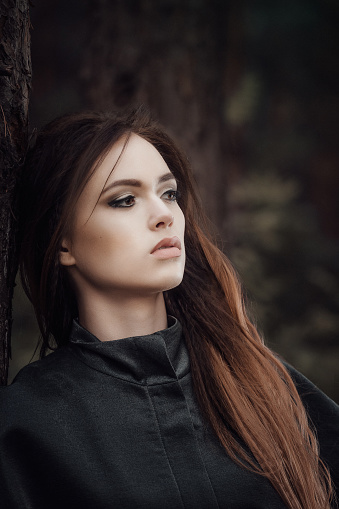 Female portrait of woman outdoors in the forest. Fairy tale atmosphere in dark. Fashion people in beautiful brown coat. Woodland dweller in the nature.